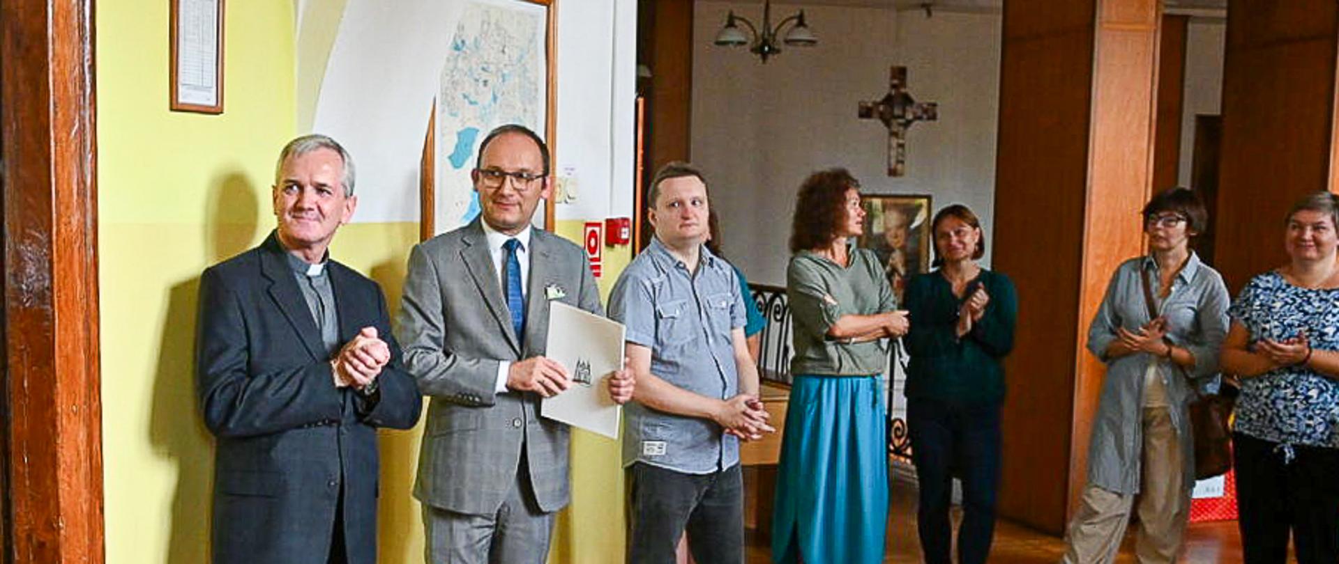 W sali z żółtymi ścianami i z podłogą z drewnianych klepek stoi kilku mężczyzn w garniturach.