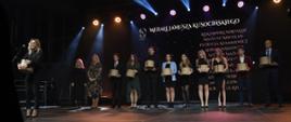 Na zdjęciu scena, a na niej kobieta mówiąca do mikrofonu, za kobietą stoi 11 osób - kobiet i mężczyzn, większość z nich trzyma ozdobne pudełka, za nimi ekran z napisami: Medale Janusza Kusocińskiego i nazwiskami.