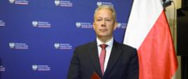Siwy mężczyzna w garniturze stoi na tle niebieskiej ściany, w rękach trzyma czerwoną teczkę, obok polska flaga.