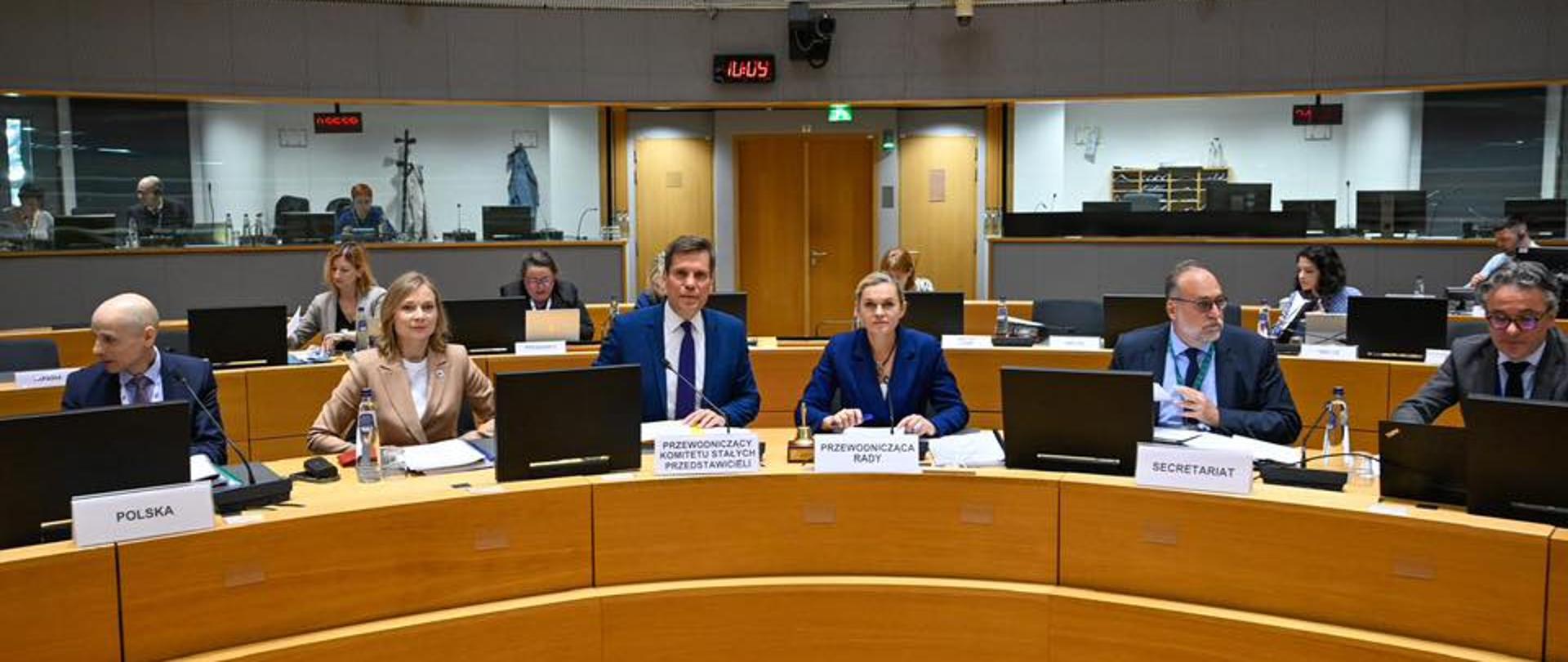 W rozległej, okrągłej sali konferencyjnej, przypominającej audytorium, siedzi grupa osób za półkolistym stołem. Na pierwszym planie widać pięcioro przedstawicieli. Po lewej stronie siedzi mężczyzna z łysą głową w ciemnym garniturze, przed którym widnieje plakietka z napisem „POLSKA”. Obok niego siedzi kobieta z jasnymi włosami w beżowej marynarce. Następnie siedzi mężczyzna w niebieskim garniturze, przed którym stoi plakietka z napisem „PRZEWODNICZĄCY KOMITETU STAŁYCH PRZEDSTAWICIELI”.
Po jego prawej stronie siedzi kobieta z blond włosami w niebieskiej marynarce, a przed nią stoi plakietka z napisem „PRZEWODNICZĄCA RADY”. Na końcu po prawej stronie siedzi mężczyzna w ciemnym garniturze i okularach, a przed nim widnieje plakietka z napisem „SECRETARIAT”.
Wszyscy przedstawiciele siedzą przy mikrofonach i laptopach, a na stołach widać dokumenty i butelki wody. W tle, na wyższych poziomach sali, siedzą inne osoby. Na ścianie w górnej części widoczny jest elektroniczny zegar wskazujący godzinę 11:09. Oświetlenie jest jasne, padające z góry. Atmosfera sugeruje formalne spotkanie Rady Unii Europejskiej