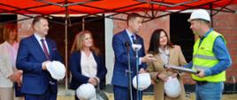 Na placu budowy, pod czerwonym namiotowym daszkiem rozpiętym na metalowym szkielecie stoją minister Czarnek, kobieta w granatowej marynarce, mężczyzna w garniturze i kobieta w jasnym szarożółtym płaszczu, trzymają w rękach białe kaski, przed mini mężczyzna w żółtej kamizelce i w kasku na głowie trzyma w rękach metalowy cylinder, w tle ściany z pomarańczowych pustaków.