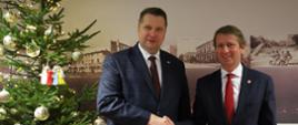 Minister Czarnek ściska rękę mężczyźnie w garniturze, obok nich choinka z bombkami, w tle ściana na której wiszą czarno-białe obrazki z różnymi budynkami.