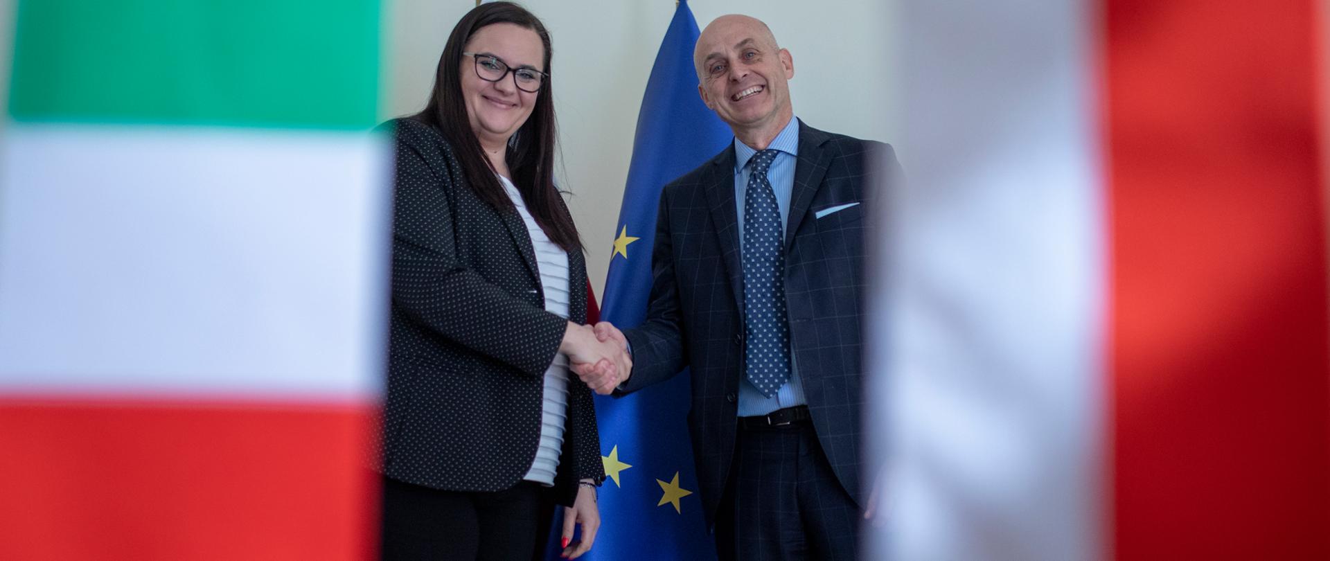 Na zdjęciu na pierwszym planie flagi Polski i Włoch. W oddali w uścisku dłoni stoją: minister Małgorzata Jarosińska-Jedynak i ambasador Włoch Aldo Amati.