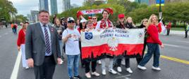 Widok na ulicę, kilkoro młodych ludzi w kolorowych strojach niesie rozpostartą polska flagę z napisem Polish Student Alliance, obok nich wiceminister Rzymkowski, w tle wysokie budynki.