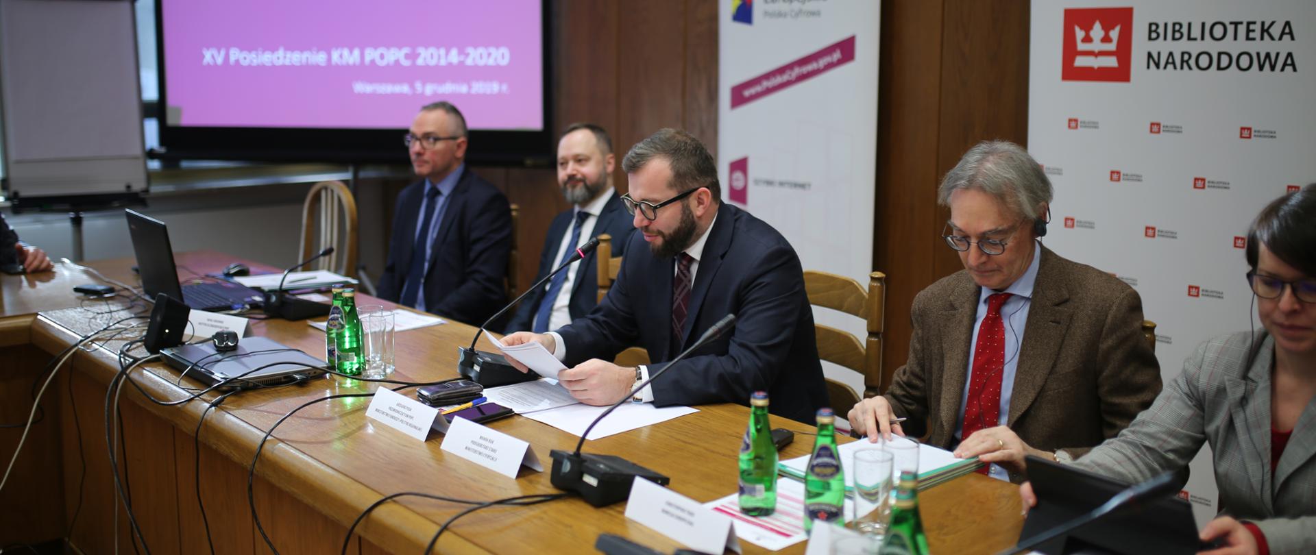 Przy długim stole siedzi pięć osób. Na stole stoją laptopy i mikrofony. Pośrodku siedzi wiceminister Grzegorz Puda. Za siedzącymi stoją banery biblioteki Narodowej i programu Polska Cyfrowa. Obok ekran.
