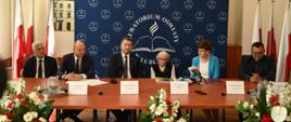 Za drewnianym stołem siedzi minister Czarnek, trzech mężczyzn, kobieta w czarnej kamizelce i kobieta w niebieskiej marynarce.