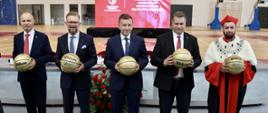 Na sali stoi w rzędzie pięciu mężczyzn, czterech w garniturach, jeden w ceremonialnym czerwonym stroju, każdy trzyma żółtą piłkę.