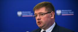 Wiceminister stoi i mówi do mikrofonu stojącego na stojaku, za nim niebieska ściana.