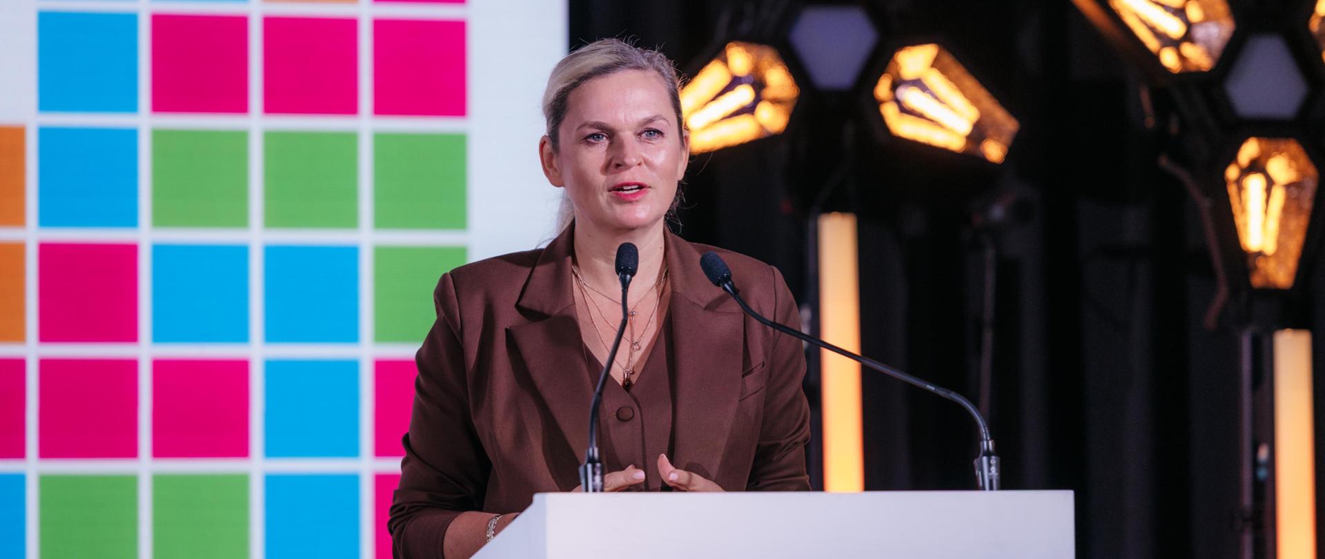 Kobieta w brązowym garniturze stoi za białym podium i przemawia do mikrofonu, patrząc z poważną miną na widownię. W tle po lewej stronie znajduje się duża, kolorowa ściana w kratkę, a po prawej widać reflektory o industrialnym wyglądzie. Na podium, na wysokości dolnej części klatki piersiowej kobiety, znajduje się ekran, na którym wyświetlony jest napis: „19. Międzynarodowa Konferencja Bezpieczeństwo dzieci i młodzieży w internecie 23-26 września 2025”.