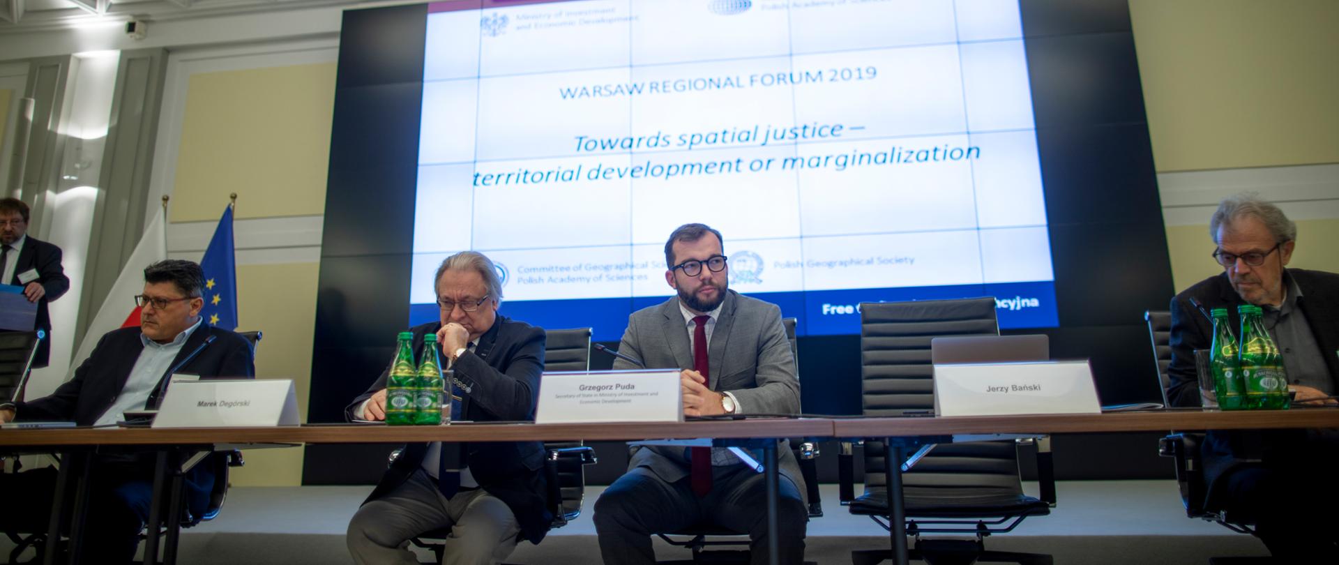 Minister Grzegorz Puda siedzi przy stole, obok niego siedzi Marek Degórski i Jerzy Bański. Za nimi na tablicy wyświetlany jest napis "Warsaw Regional Forum 2019. Towards spatial justice - territorial development or marginalization"