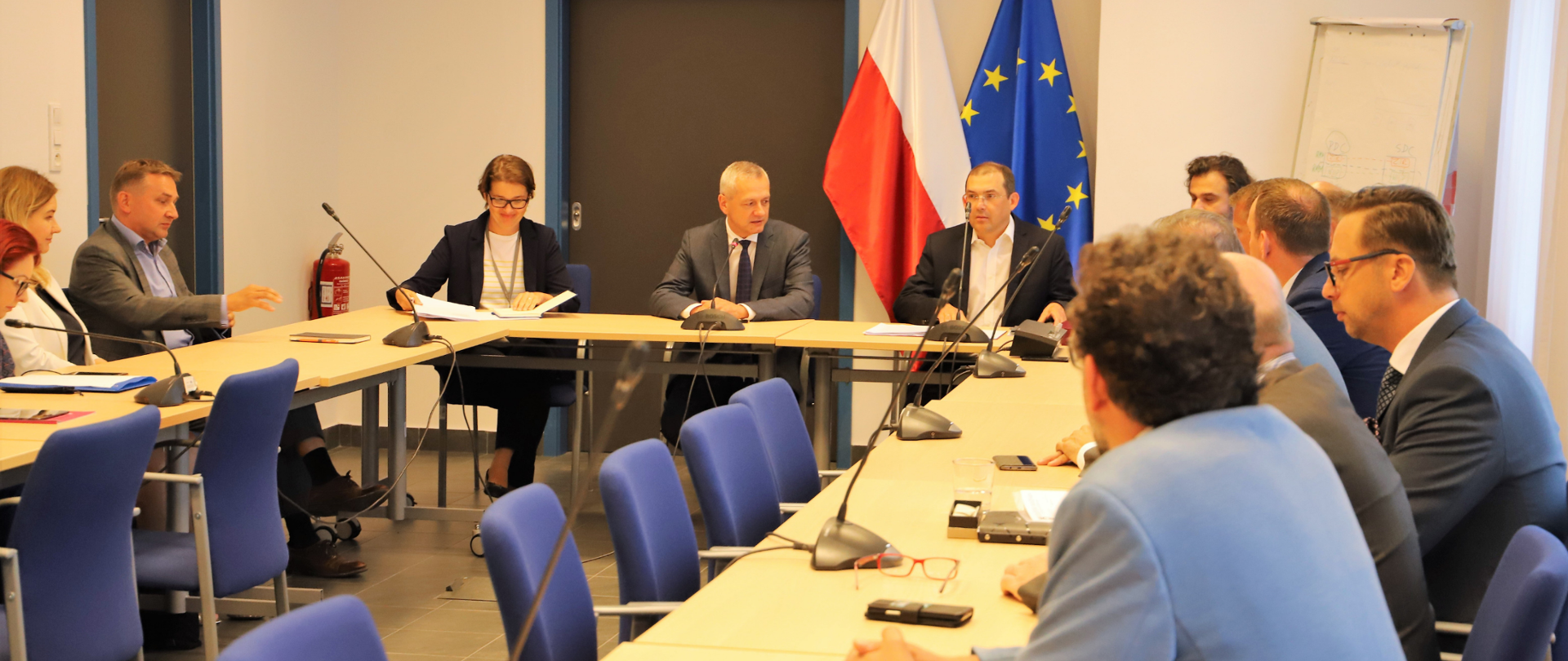 Sala konferencyjna. Kilkanaście osób siedzi przy stolikach. Na końcu sali siedzi minister Marek Zagórski i pracownicy ministerstwa. W tle flagi: polska i Unii Europejskiej.