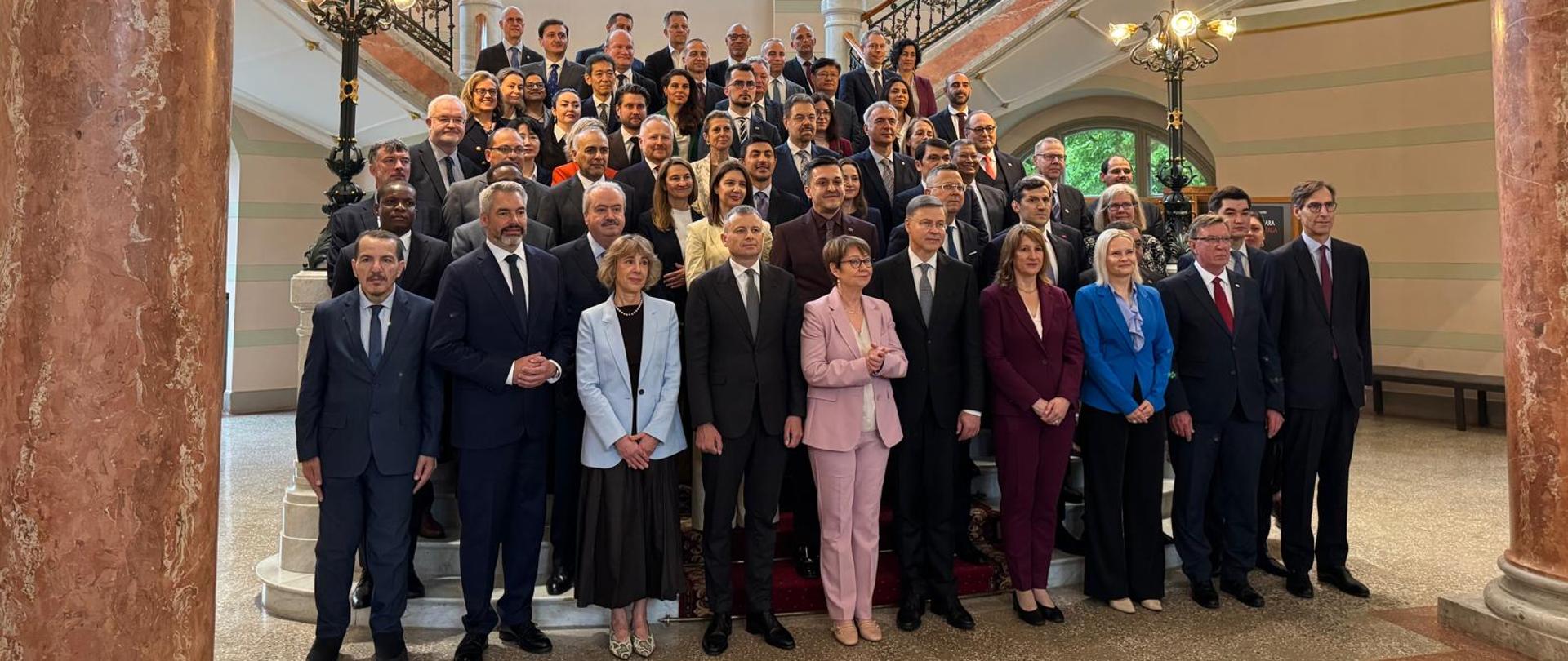 Duża grupa osób w formalnych strojach stoi na schodach ozdobnego wnętrza z kolumnami, wysokimi oknami i dekoracyjnymi balustradami, pozując do wspólnego zdjęcia.