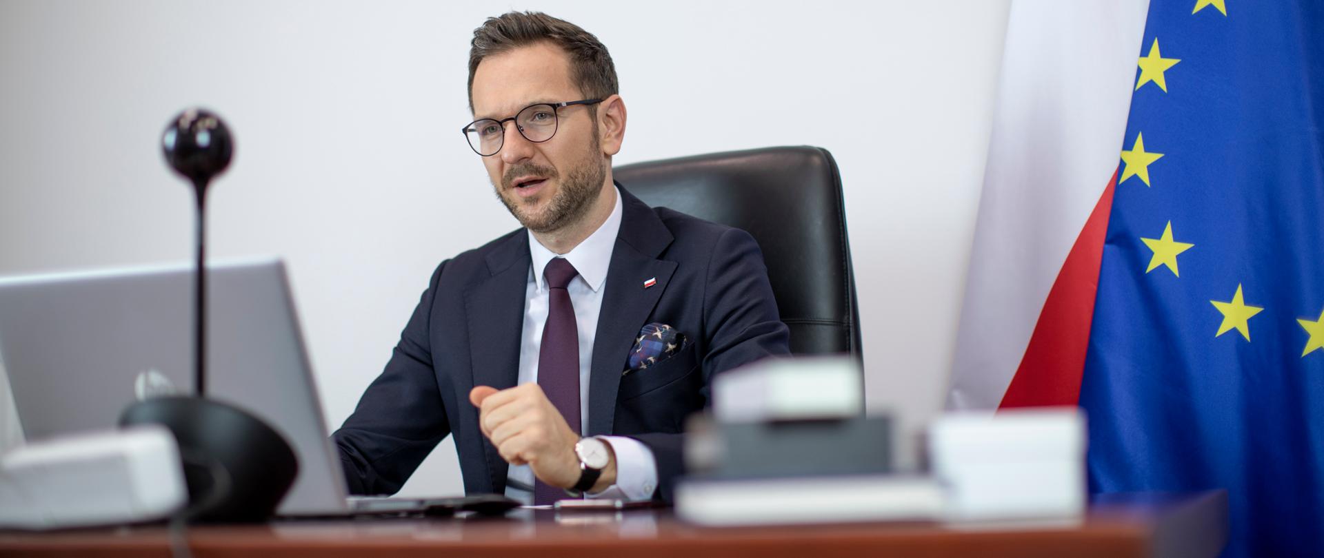 wiceminister Waldemar Buda przed komputerem, za nim flagi Polski i Unii Europejskiej