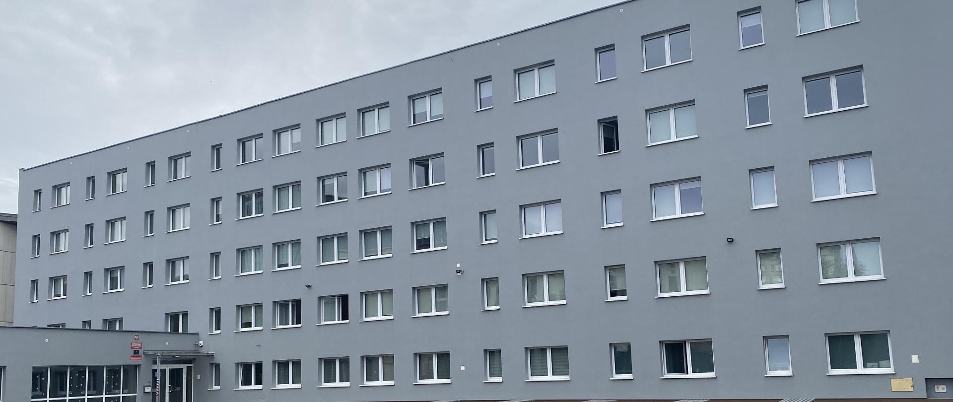 Zdjęcie przedstawia popielaty budynek bursy z licznymi oknami, po lewj stronie znajdują się drzwi wejściowe, przed budynkiem jest pięć ławek
