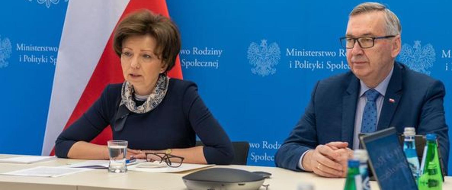 Za stołem siedzą minister Maląg i wiceminister Szwed. Z tyłu widoczny niebieski baner z logo Ministerstwa Rodziny i Polityki Społecznej. Na stole stoją butelki z wodą. Przed minister Maląg napełniona wodą szklanka, obok okulary.