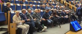 Widok z boku na salę, na krzesłach siedzą ludzie.