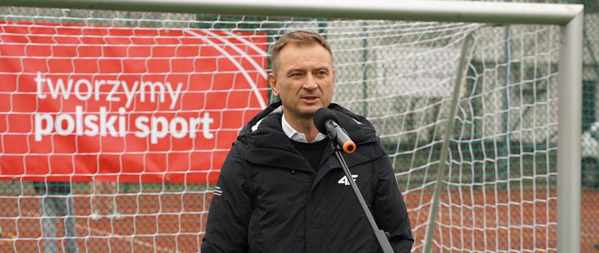 Podsumowanie 100 dni pracy Ministerstwa Sportu i Turystyki - na zdjęciu minister Sławomir Nitras mówi do stojącego przed nim mikrofonu, w tle bramka do piłki nożnej i baner z napisem "tworzymy polski sport"