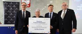 Trzech mężczyzn w garniturach i trzymająca dużą kartę z napisem Branżowe Centrum Umiejętności - 14 596 474,64 zł kobieta stoją w rzędzie.
