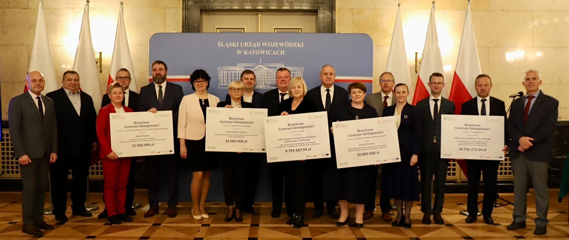 Na sali z mozaikową podłogą i żółtymi marmurowymi ścianami stoi w rzędzie kilkanaście osób, pięć trzyma białe tabliczki z napisami Branżowe Centrum Umiejętności.