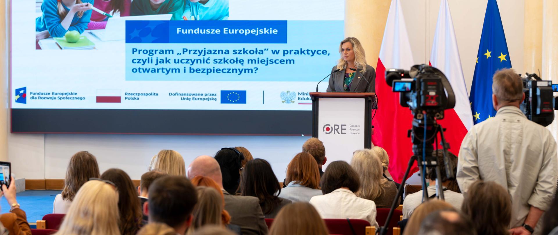 Szerokie ujęcie sali konferencyjnej. Blondynka przemawia przy mownicy ORE, a obok stoją flagi Polski i UE. W tle widoczny jest duży ekran z prezentacją i tekstem: „Program «Przyjazna szkoła» w praktyce...”. Na pierwszym planie widać siedzącą publiczność oraz kamery telewizyjne.