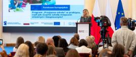 Szerokie ujęcie sali konferencyjnej. Blondynka przemawia przy mownicy ORE, a obok stoją flagi Polski i UE. W tle widoczny jest duży ekran z prezentacją i tekstem: „Program «Przyjazna szkoła» w praktyce...”. Na pierwszym planie widać siedzącą publiczność oraz kamery telewizyjne.