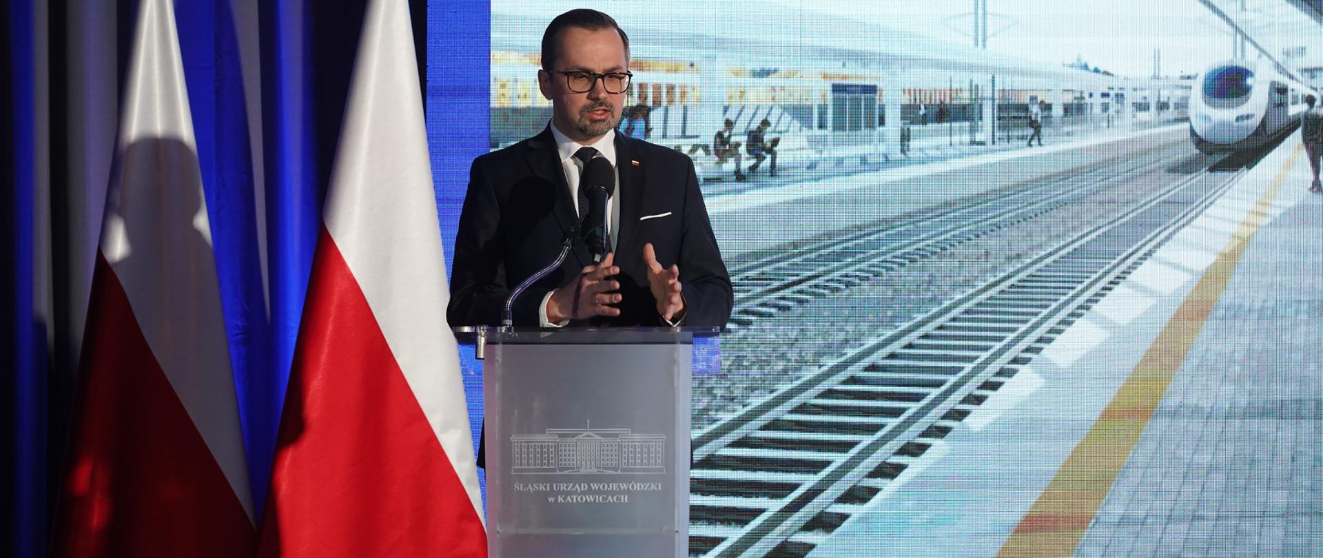 Wiceminister Marcin Horała stoi na scenie przy pulpicie i mówi do mikrofonu. 