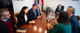 Grupa kilku osób siedzi dookoła owalnego stołu i rozmawia, za nimi na białej ścianie wiszą kolorowe obrazki.