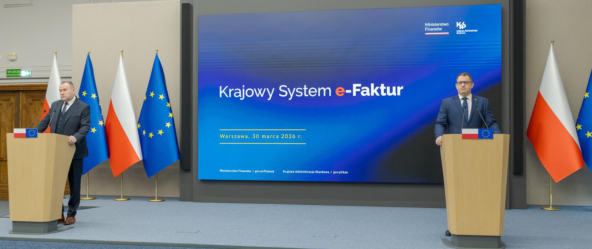 Marcin Łoboda szef Krajowej Administracji Skarbowej oraz Zbigniew Stawicki zastępca szefa KAS podczas konferencji prasowej w Ministerstwie Finansów opowiedzieli o wdrożeniu II etapu Krajowego Systemu e-Faktur przy mównicach przed dużym ekranem z napisem „Krajowy System e‑Faktur”, obok flag Polski i Unii Europejskiej.