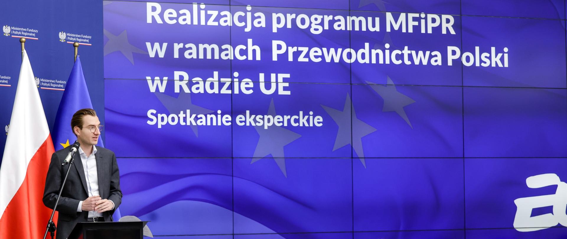 Wiceminister Jan Szyszko stoi z lewej strony przy pulpicie u mówi do osób zgromadzonych na sali. 
