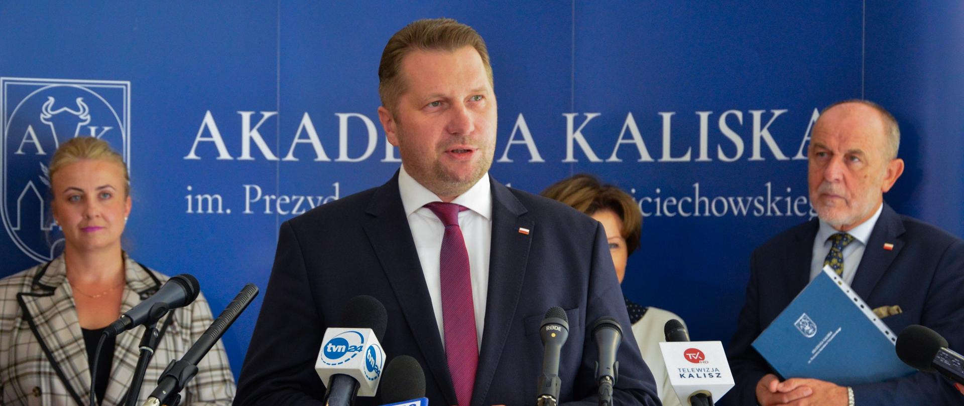 Minister w otoczeniu kilku osób stoi i mówi do ustawionego na stojaku mikrofonu, za nim niebieska ściana z napisem Akademia Kaliska.