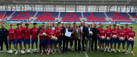 Zbiorowe zdjęcie przedstawiające wiceministra sportu i turystyki Piotra Borysa z członkami Akademii Odry Opole. Zdjęcie zrobione jest na murawie stadionu. W tle widać trybuny z napisem "Opole".