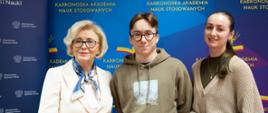 Wiceminister Machałek w białym ubraniu stoi na tle niebieskiej ścianki z napisem Karkonoska Akademia Nauk Stosowanych, obok niej młody chłopak w szaro-brązowej bluzie z kapturem i dziewczyna w sweterku.