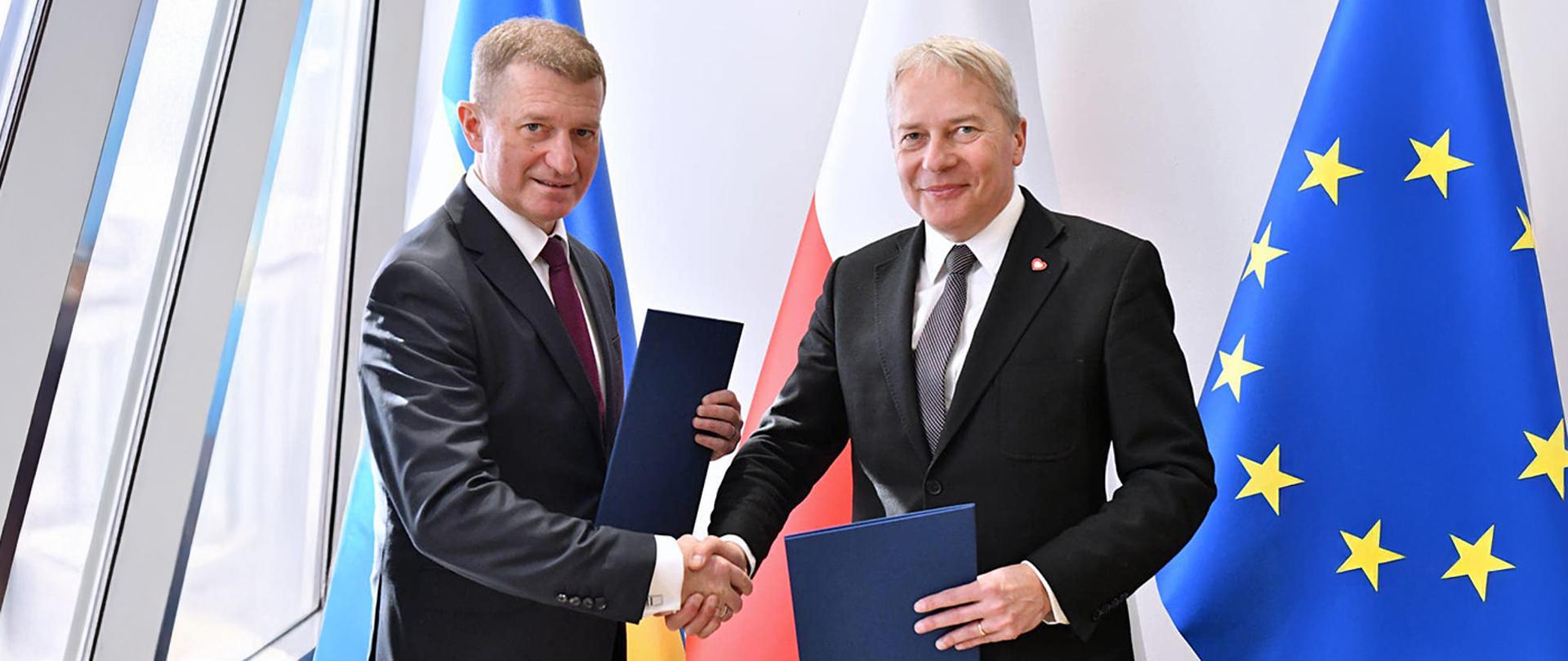 Zdjęcie Sekretarza Stanu w MSiT Ireneusza Rasia, który ściska dłoń Marszałka Województwa Śląskiego Wojciecha Saługi. Panowie trzymają teczki, w których znajdują się podpisane porozumienia w sprawie współpracy na rzecz realizacji Programu rozwoju infrastruktury sportowej w województwach. W tle flagi Polski i Unii Europejskiej.