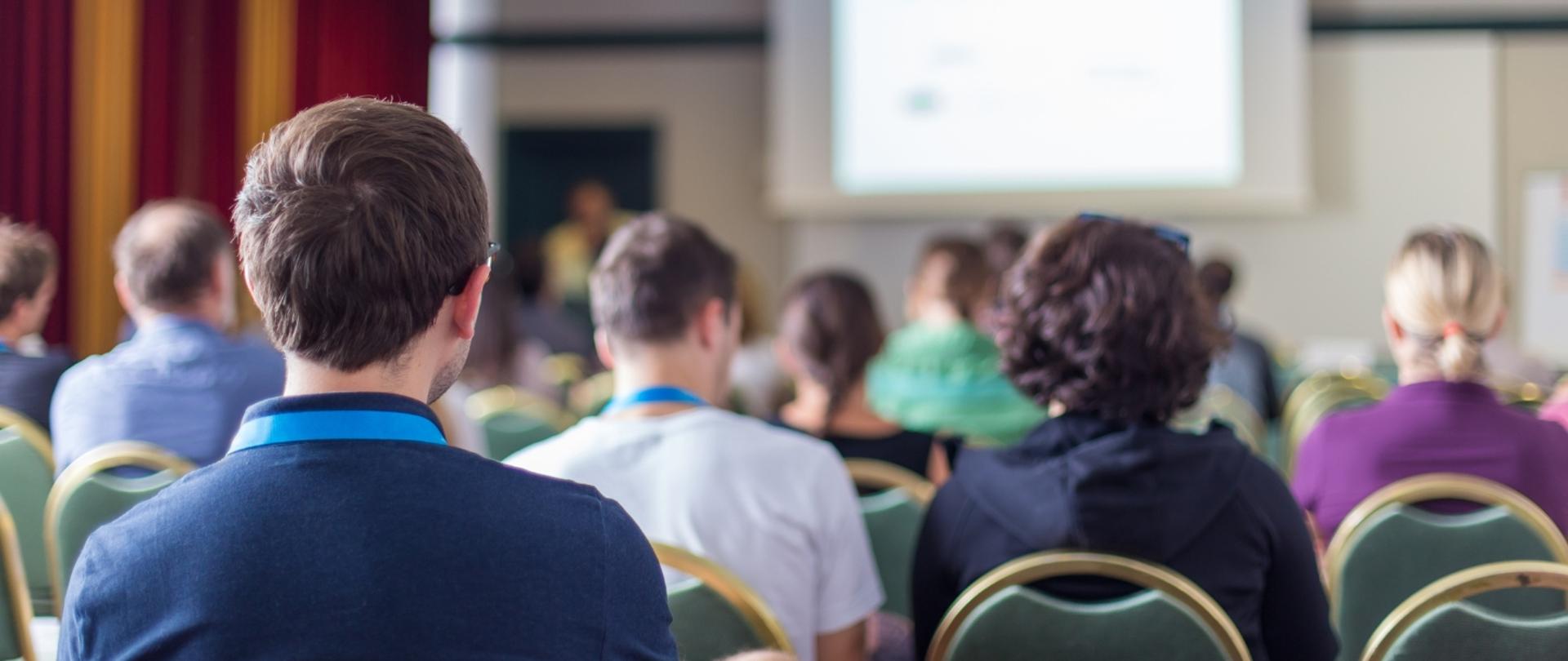 na zdjęciu widnieją ludzie uczestniczący w konferencji, przed nimi znajduje się rzutnik 