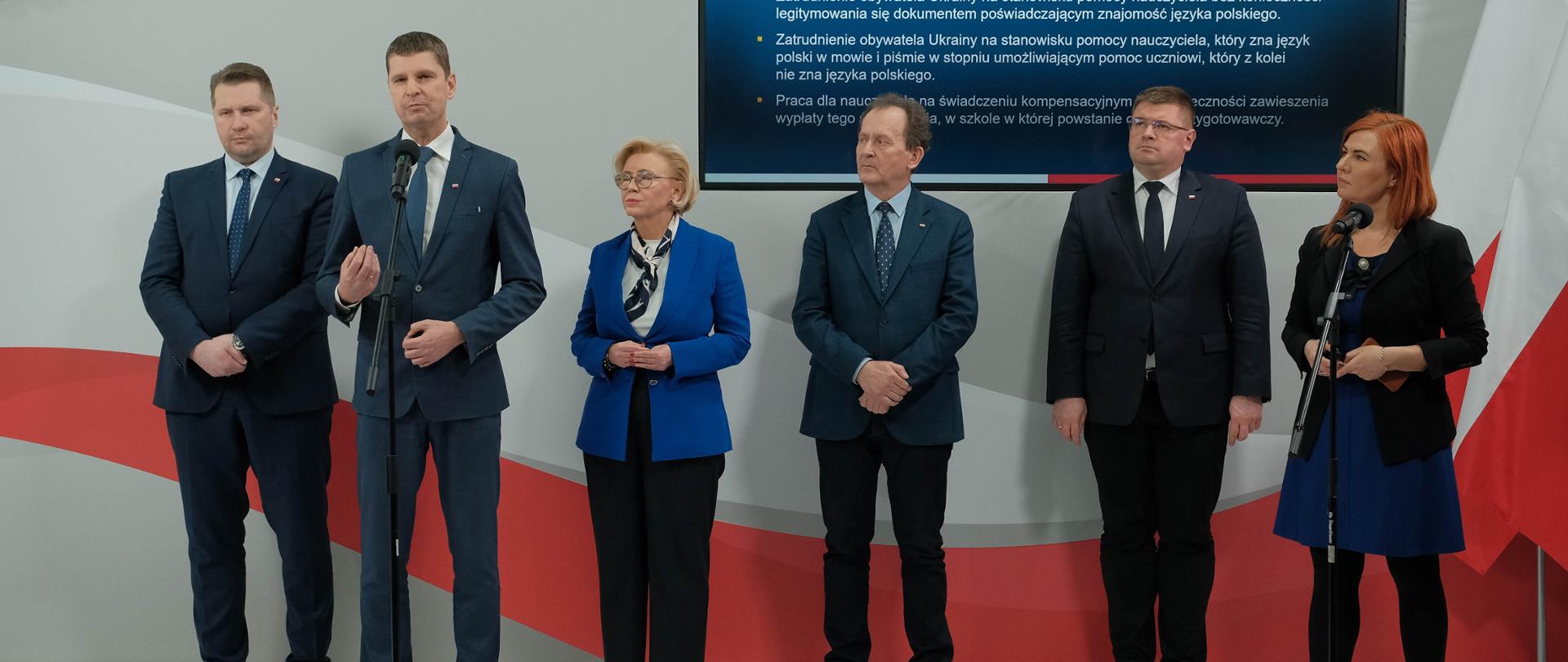 Od lewej minister Przemysław Czarnek, wiceminister Dariusz Piontkowski, wiceminister Marzena Machałek, wiceminister Włodzimierz Bernacki, wiceminister Tomasz Rzymkowski oraz rzecznik prasowy Anna Ostrowska. Wiceminister Piontkowski stoi przy mikrofonie. Biało-czerwone tło w tle oraz ekran z tekstem. 