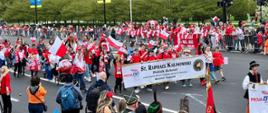 Widok z góry i z boku na wypełnioną niosącymi polskie flagi ludźmi ulicę, w tle zielone drzewa i wysokie budynki.