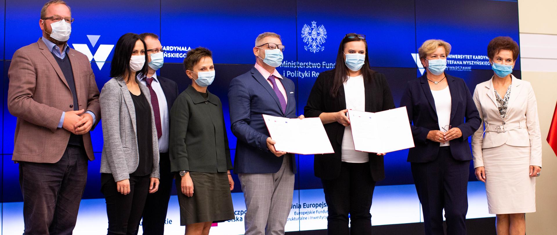 Na scenie stoi grupa ludzi. Dwie z nich trzymają w rękach otwarte dokumenty. Za nimi na ekranie granatowe tło.
