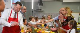 Grupa małych dzieci w białych fartuszkach stoi dookoła stołu, na którym jest dużo kolorowych owoców i miski z różnokolorowymi płynami, przy nich kilkoro dorosłych w czerwonych fartuchach.