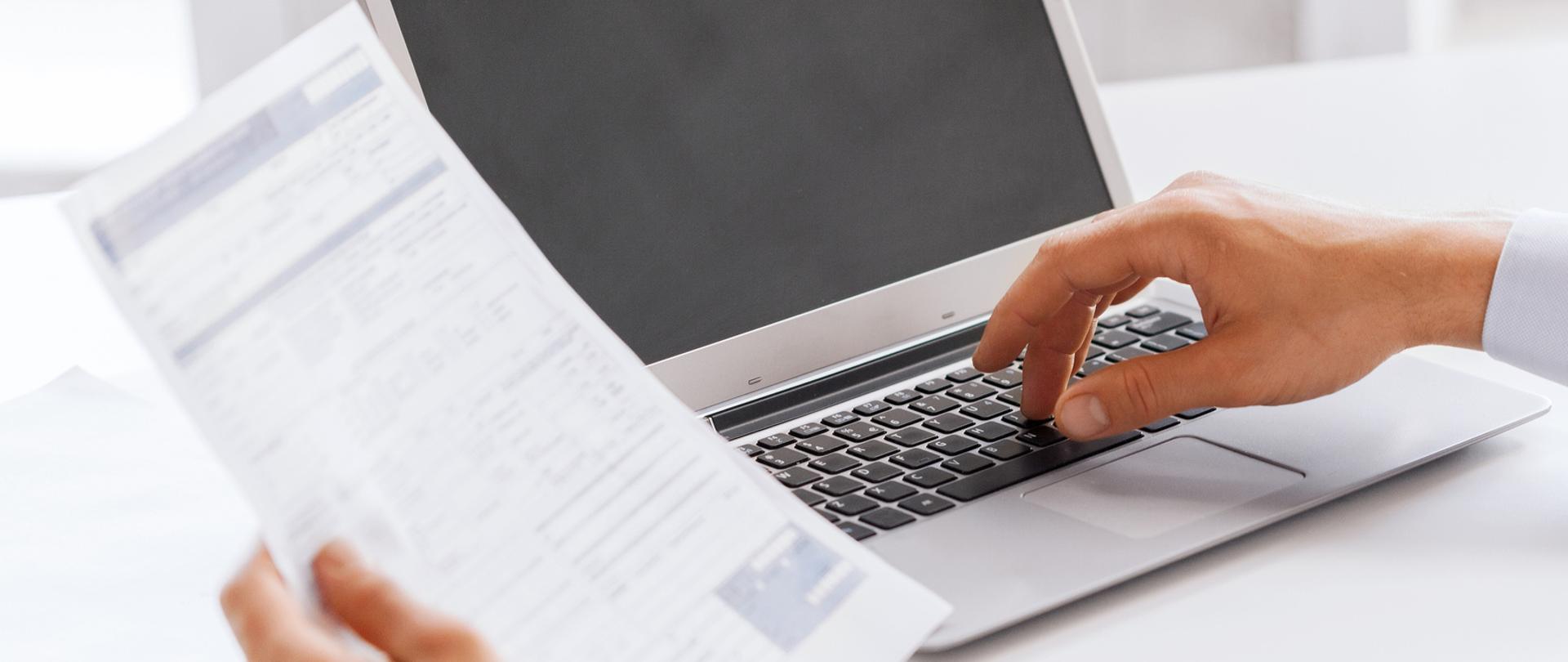 Biały laptop na tle białego blatu stołu i dwie dłonie w których w jednej widać formularz.