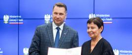 Minister Czarnek i ubrana na czarno kobieta stoją obok siebie, razem trzymają otwartą teczkę z dokumentami.