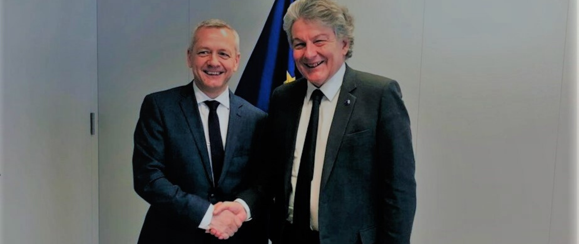 Minister Marek Zagórski i unijny komisarz Thierry Breton pozują do zdjęcia ściskając sobie dłonie na tle unijnej flagi
