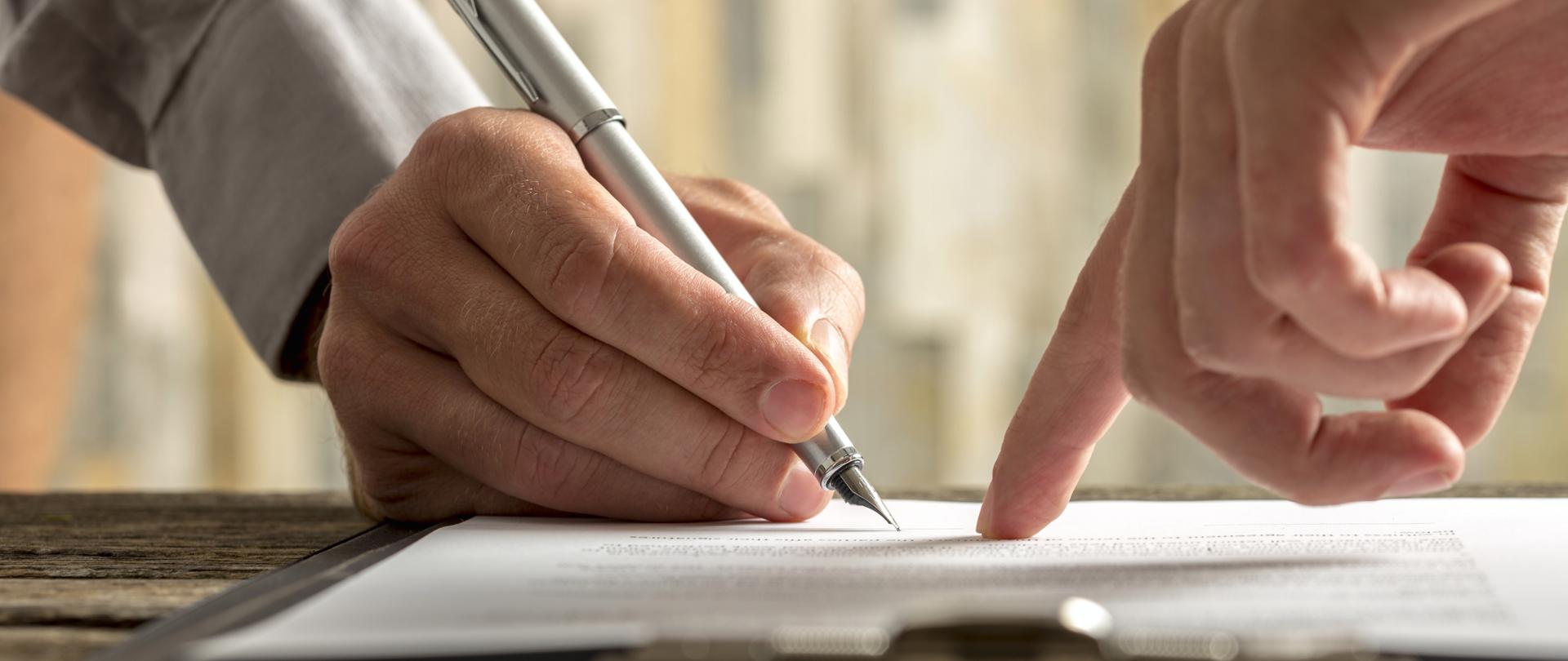 Karta papieru a nad nią dwie dłonie jedna wskazuje palcem a druga dotyka wiecznym piórem.