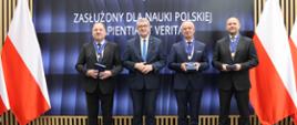 Pod wbudowanym w ścianę wielkie ekranem z napisem Ceremonia wręczenia medali „Zasłużony dla Nauki Polskiej Sapientia et Veritas” stoi czterech mężczyzn w garniturach, po bokach polskie flagi.
