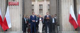 Przed wejściem do szarego budynku stoi grupa ludzi, na ścianie tabliczka Ministerstwo Edukacji i Nauki, po bokach wejścia polskie flagi.