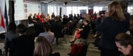 sala wypełniona uczestnikami konferencji, w oddali widoczne na scenie panelistki