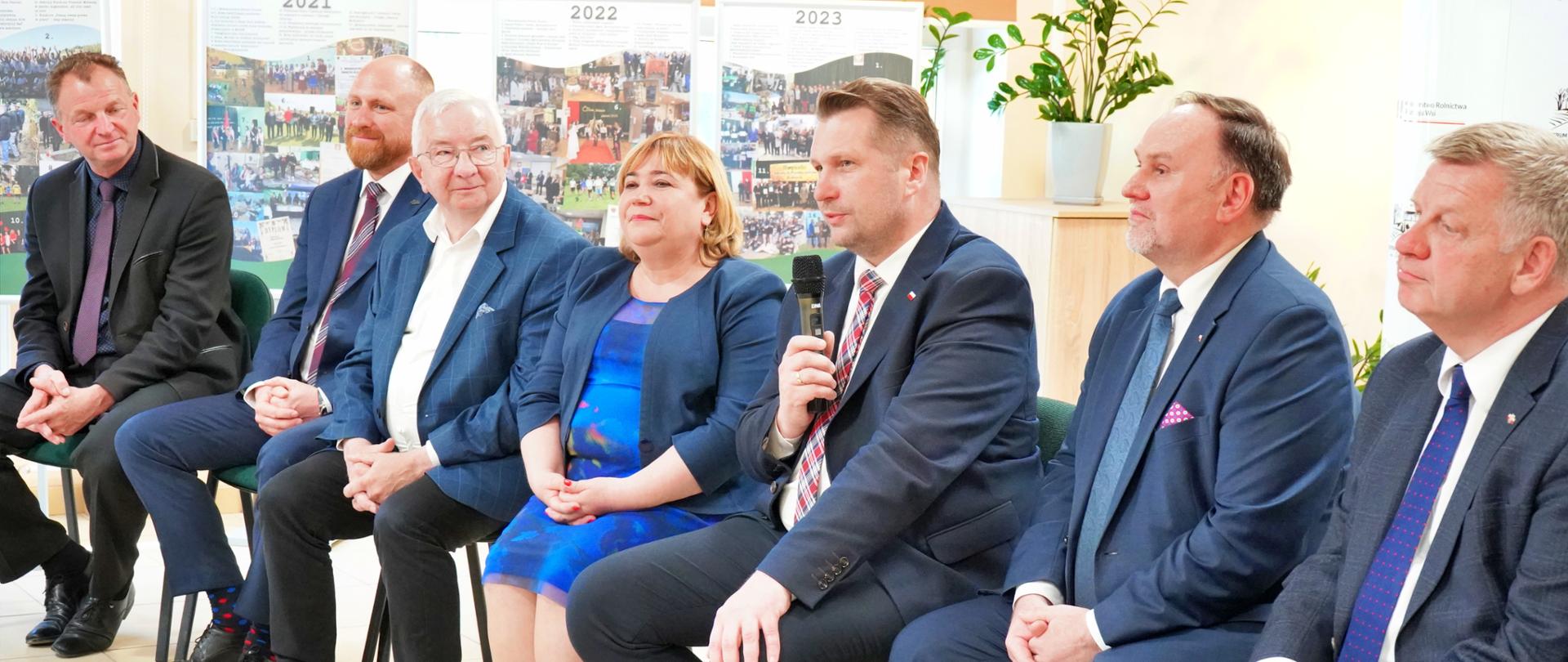 Na ustawionych w rzędzie krzesłach siedzi siedem osób, pośrodku minister Czarnek mówi do trzymanego w ręku bezprzewodowego mikrofonu, w tle żółta ściana na której wiszą korkowe tablice z przypiętymi kolorowymi kartkami.