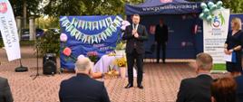 Na wyłożonym szaro-czerwoną kostką placyku stoi minister Czarnek i mówi do mikrofonu, za nim niebieski namiotowy daszek z napisem www.powiatleczynski.pl, rozwieszony niebieski materiał z napisem Piknik integracyjny i zielone drzewa, przed nim na krzesłach siedzą ludzie.