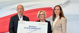 Wiceminister Machałek stoi obok mężczyzny w garniturze i kobiety w beżowym ubraniu, wszyscy trzymają wielki symboliczny czek z napisem Branżowe Centrum Umiejętności - 15 000 000 zł.
