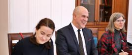 Przy drewnianym stole siedzi uśmiechnięty mężczyzna w garniturze, po obu stronach dwie kobiety, które piszą w zeszytach - ubrana na czarno i w marynarce w czerwono-czarną kratę, za nimi biała ściana i drewniana szafka z książkami.