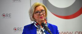 Minister Machałek w niebieskim ubraniu stoi i mówi do mikrofonu, za nią ściana z napisami ORE.